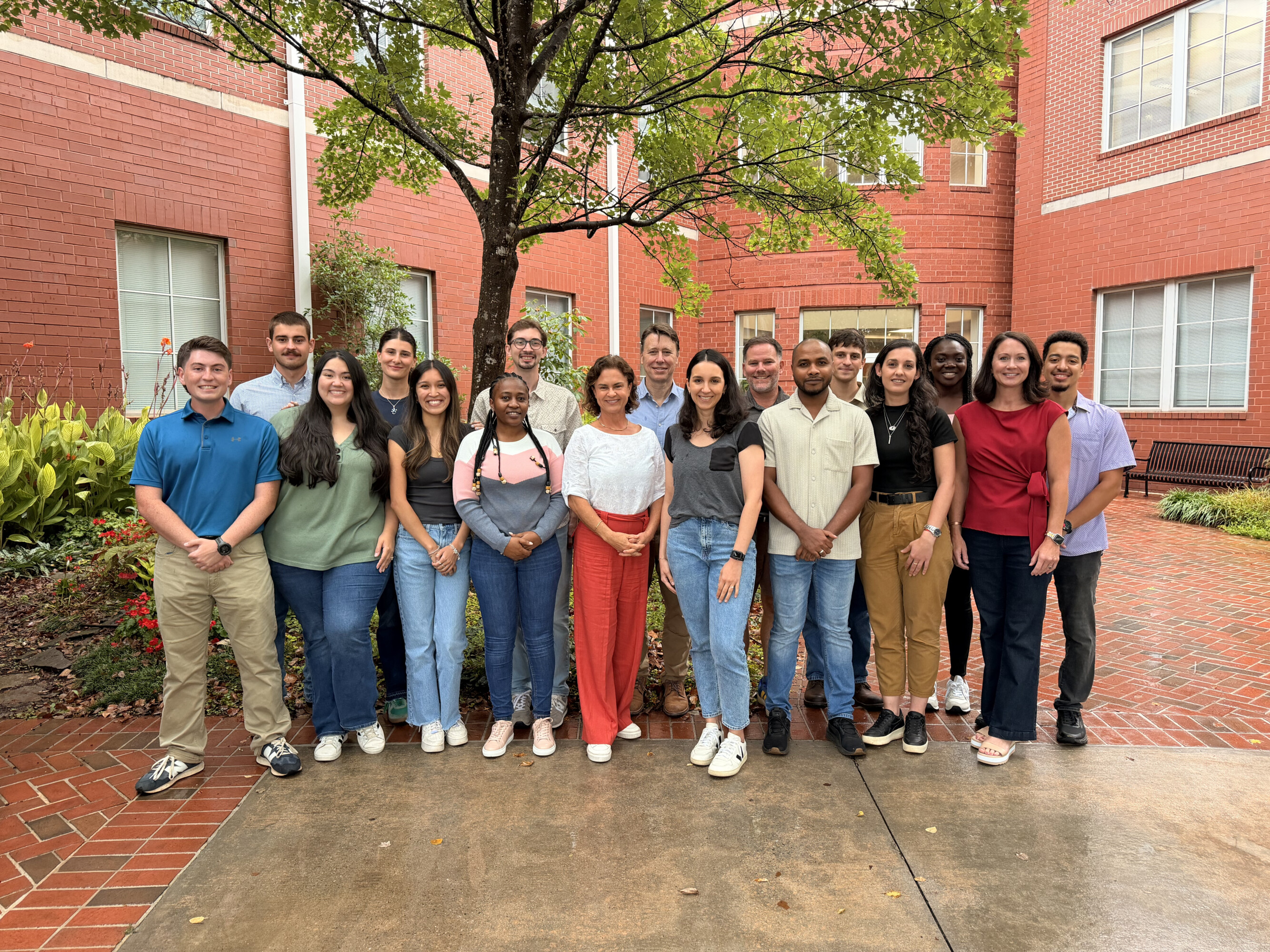 A formal group portrait of the Wild Peanut Lab team standing outside the Center for Applied Genetic Technologies (CAGT) at the University of Georgia. The researchers are posed together in front of the building’s red brick exterior, framed by landscaped flower beds. This image serves as the lab's primary team photo for the 2026 academic year.